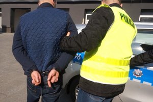 Na zdjęciu widać policjanta w żółtej kamizelce odblaskowej, który wyprowadza do radiowozu podejrzanego z zapiętymi z tyłu kajdankami.