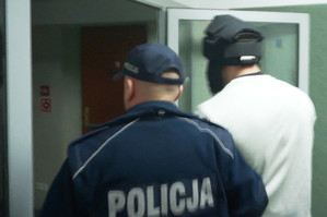 Zdjęcie policjanta prowadzącego zatrzymanego mężczyznę  w jasnej bluzie i ze specjalnym kaskiem ochronnym na głowie.