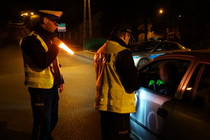W nocy  na drodze dwóch policjantów z Wydziału Ruchu Drogowego kontroluje trzeźwość kierowcy. Jeden z nich oświetla miejsce kontroli latarką.