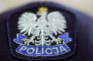Zdjęcie emblematu mundurowego w postaci wyhaftowanej naszywki na czapce przedstawiającej ukoronowanego białego orła na granatowej tarczy. Poniżej napis „policja”.