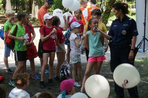 Dziewczynka udzielająca do mikrofonu odpowiedzi policjantce, za nimi grupka dzieci z opiekunem. Niektóre dzieci trzymają białe balony.