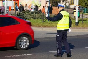 Policjant na skrzyżowaniu kieruje ruchem. Przed policjantem przejeżdża czerwony samochód osobowy.