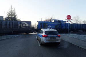 Policyjny radiowóz stojący przed przejazdem kolejowym na którym zatrzymały się dwie lokomotywy.