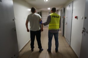 Na korytarzu widoczny policjant w żółtej kamizelce z napisem POLICJA, prowadzi zatrzymanego mężczyznę.