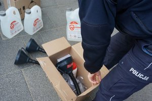 Częściowo widoczny policjant, pakujący do kartonowego pudełka środki ochronne. Widoczne środki ochronne - maseczki, płyny do dezynfekcji, lejki.
