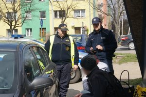 Policjant i strażnik miejski legitymujące siedzące osoby. Widoczne częściowo samochody, w tle blok mieszkalny i przechodnie.