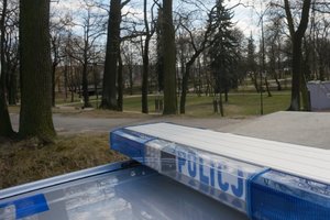 Widoczny częściowo radiowóz - &quot;koguty policyjne&quot; z napisem POLICJA. W tle widoczny opustoszały park.