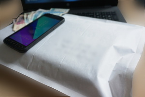 Widoczna przesyłka listowa w białej kopercie, na kórej leży czarny telefon komórkowy, a także banknoty. W tle widoczny częściowo ekran laptopa.