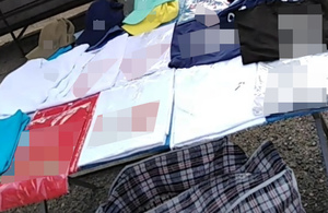 Na zdjęciu widoczne stoisko z rozłożoną do sprzedaży podrobioną odzieżą: koszulki w foliach, czapki z daszkiem. Pod stolikiem widoczna częściowo torba.