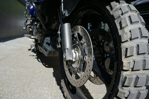 Częsciowo widoczny motocykl - na pierwszym planie przednie koło motocyklowe, w tle dalsza część pojazdu.