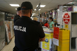 Umundurowani policjanci podczas kontroli przestrzegania przez klientów sklepu obowiązku zasłaniania nosa i ust. W tle widoczna ekspedientka sklepu i klienci.