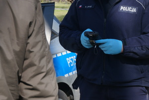 Zbliżenie na ręce umundurowanego policjanta podczas legitymowania osoby. Po lewej stronie widoczna częściowo osoba. W tle widoczny częściowo radiowóz.
