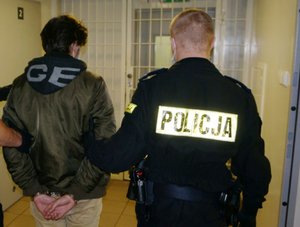 Na zdjęciu widoczny mężczyzna z założonymi na ręce trzymane z tyłu kajdankami, prowadzony przez umundurowanego policjanta.