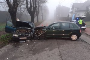 Na zdjęciu widoczne dwa zderzone ze sobą samochody z uszkodzonymi przodami. Po prawej stornie widoczny umundurowany policjant w trakcie czynności. Po lewej stronie za pojazdem widoczna częściowo osoba.