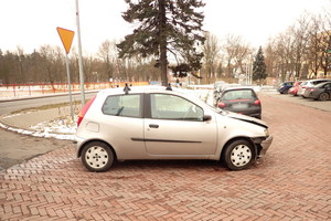 Na zdjęciu widoczny bokiem samochód osobowy z roztrzaskanym z prawej strony błotnikiem i maską. Po lewej stronie widoczny pionowy znak drogowy - ustąp pierwszeństwa przejazdu. W tle widoczne zaparkowane samochody.