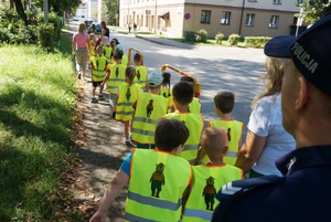Na zdjęciu widoczne dzieci w kamizelkach odblaskowych idące chodnikiem oraz częściowo widoczny policjant.