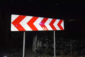 Na zdjęciu widoczne oznakowanie pionowe prowadzące w lewo, za którym widoczny jest przewrócony samochód ciężarowy.