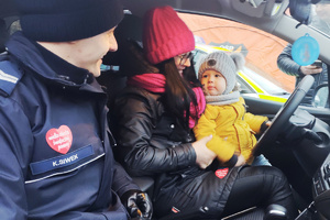 Na zdjęciu widoczny policjant siedzący w radiowozie, a za kierownicą kobieta trzymająca dziecko.