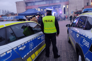 Na zdjęciu widoczny policjant, który stoi pomiędzy radiowozami i obserwuje imprezę - 32. Finał Wielkiej Orkiestry Świątecznej Pomocy.