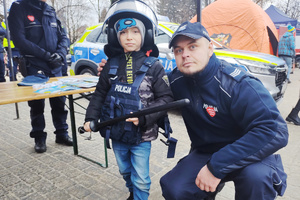 Na zdjęciu widoczny policjant pozujący do zdjęcia z chłopcem, który ma na sobie policyjny sprzęt.