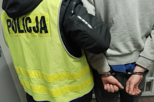 Na zdjęciu widoczny nieumundurowany policjant w kamizelce odblaskowej z napisem Policja, który prowadzi mężczyznę korytarzem. Mężczyzna na ręce trzymane z tyłu ma założone kajdanki.
