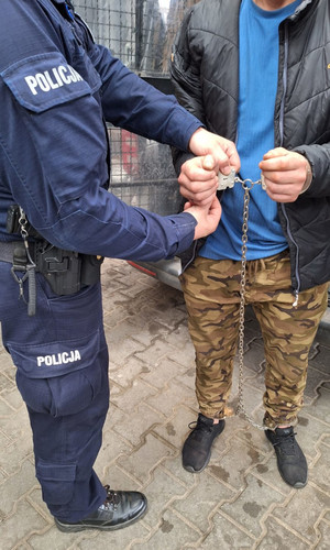 Na zdjęciu częściowo widoczny policjant, który zapina kajdanki zespolone na rękach zatrzymanego mężczyzny.